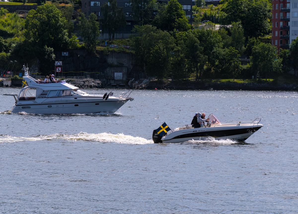 Foto eines Motorboots und einer kleinen Jacht im Meer rund um Stockholm. Im Hintergrund sind Bäume, Gebäude und Straßen.
