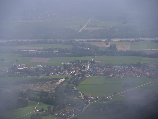 Bild eines kleinen Dorfes in Bayern, Deutschland. Das Bild wurde von einem Berg aus durch den Nebel aufgenommen. Hinter dem Dorf sind im Hintergrund ein Fluss und ein weiteres Dorf zu sehen.