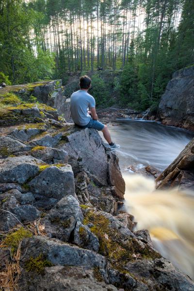 Zu sehen ist ein Wasserfall mit verwischtem Wasser und einigen Felsen zur Linken. Auf diesen Felsen sitzt ein Mann, der aufs Wasser hinabblickt.