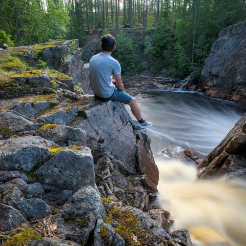 Zu sehen ist ein Wasserfall mit verwischtem Wasser und einigen Felsen zur Linken. Auf diesen Felsen sitzt ein Mann, der aufs Wasser hinabblickt.