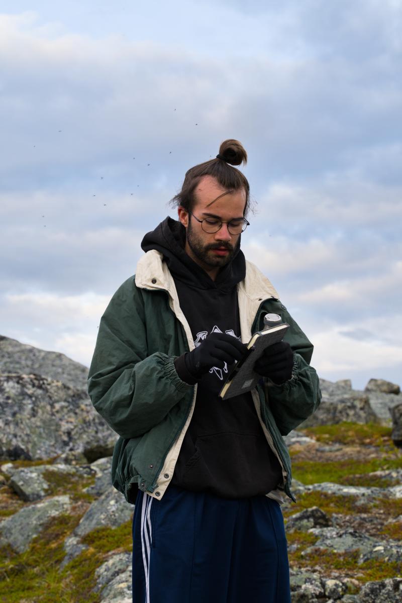 Ein Mann mit Brille und Handschuhen blickt auf ein kleines Notizbuch in seinen Händen, er trägt einen schwarzen Hoodie unter einer offenen grünen Jacke. Im Hintergrund liegen die flechtenbewachsenen Felsen und das Moos des schwedischen Fjälls unter einem bewölkten Himmel.