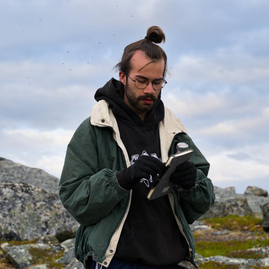 Ein Mann mit Brille und Handschuhen blickt auf ein kleines Notizbuch in seinen Händen, er trägt einen schwarzen Hoodie unter einer offenen grünen Jacke. Im Hintergrund liegen die flechtenbewachsenen Felsen und das Moos des schwedischen Fjälls unter einem bewölkten Himmel.