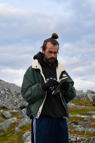 Ein Mann mit Brille und Handschuhen blickt auf ein kleines Notizbuch in seinen Händen, er trägt einen schwarzen Hoodie unter einer offenen grünen Jacke. Im Hintergrund liegen die flechtenbewachsenen Felsen und das Moos des schwedischen Fjälls unter einem bewölkten Himmel.