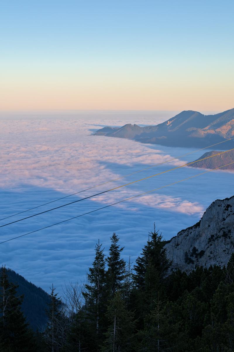 Dieses Bild wurde von oberhalb der Wolken aufgenommen. Man blickt von einem Berg auf sie hinab. Im Bild ist ein weiterer Berg zu sehen. Der Himmel ist hellblau / orange.